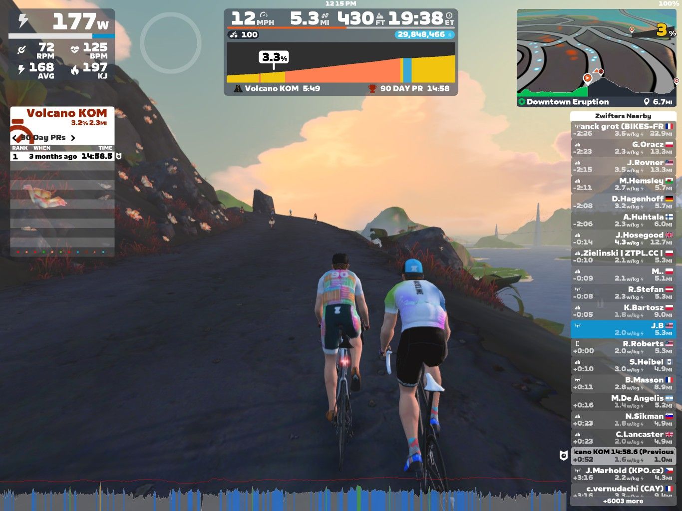 Two cyclists are riding on a virtual road in Zwift, with a scenic background featuring hills and a colorful sky. The cyclists are approximately shoulder to shoulder, and various on-screen stats display their speed, power output, and other performance metrics. The setting includes a view of a mountain labeled 