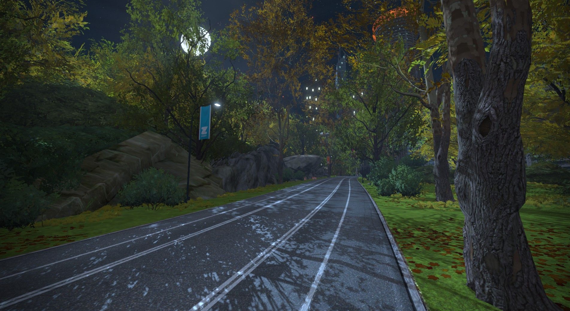 A winding path in a lush, green park is illuminated by soft lighting at night, flanked by trees with autumn foliage. A few large rocks are visible alongside the path, and a flag with the Zwift logo can be seen on a nearby pole.
