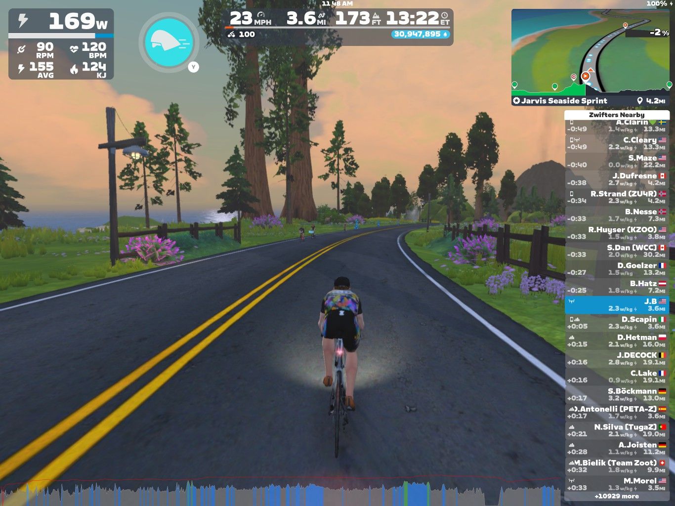 A cyclist rides on a virtual road surrounded by greenery and trees in Zwift, showcasing stats like power, speed, and distance on the screen. In the background, other cyclists can be seen along the road. The scene is set during a colorful sunrise or sunset.