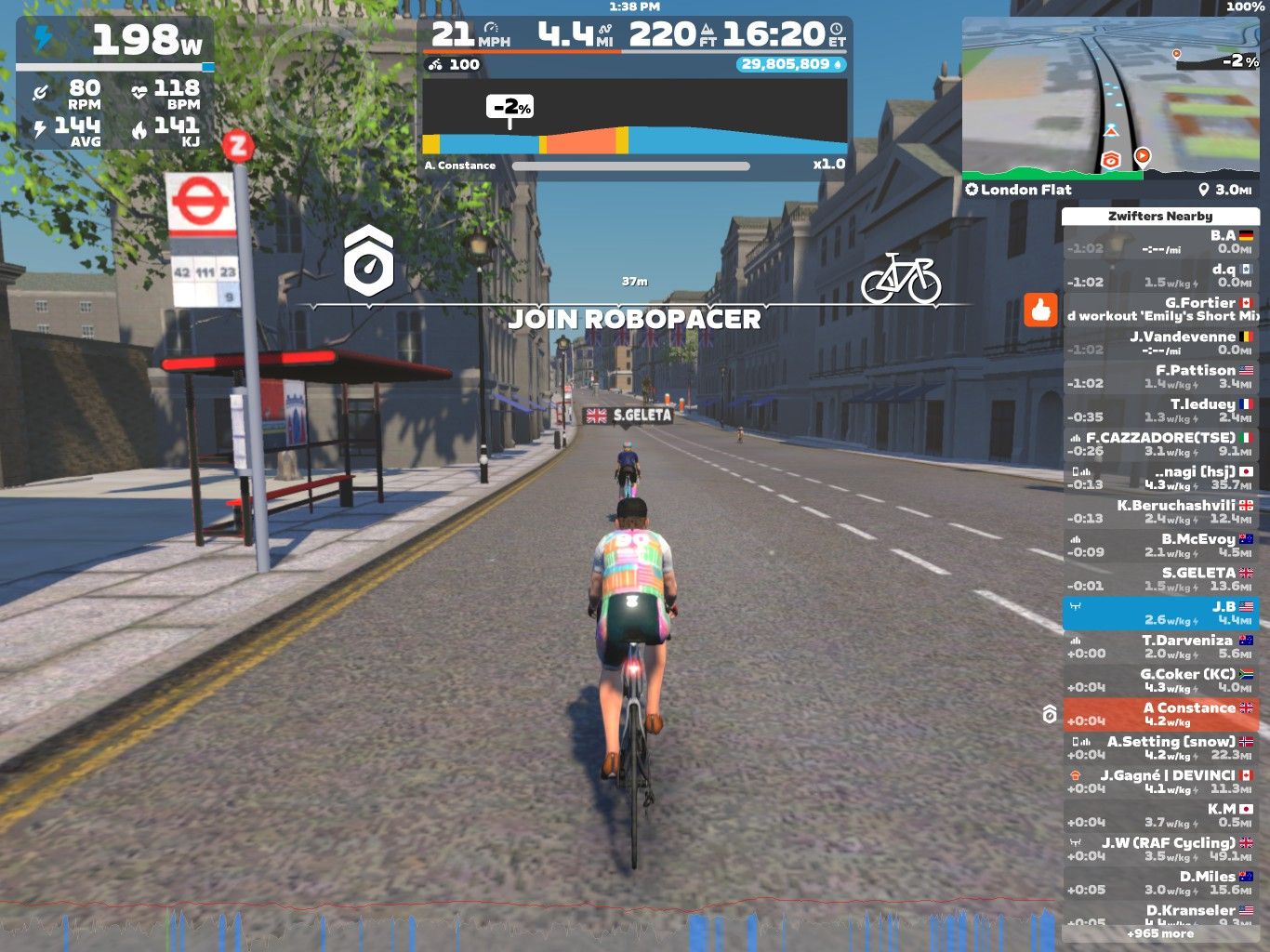 A cyclist riding on a virtual street in Zwift, with a bus stop nearby. The screen displays real-time stats such as wattage, speed, distance, and heart rate, along with a sidebar showing other users and their performance metrics. Bright sunlight and trees line the street, creating a lively outdoor atmosphere.