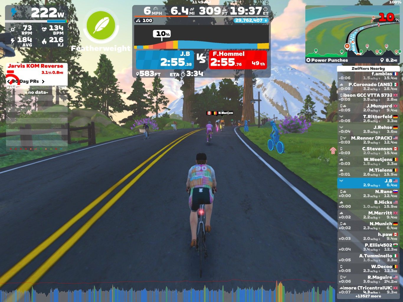 A virtual cycling scene in Zwift, showing a cyclist on a road surrounded by trees and mountains. The cyclist is riding uphill with a visible power output of 222W. On the screen, there are statistics for distance, speed, heart rate, and nearby riders. The environment features colorful graphics with flowers and a bright sky.