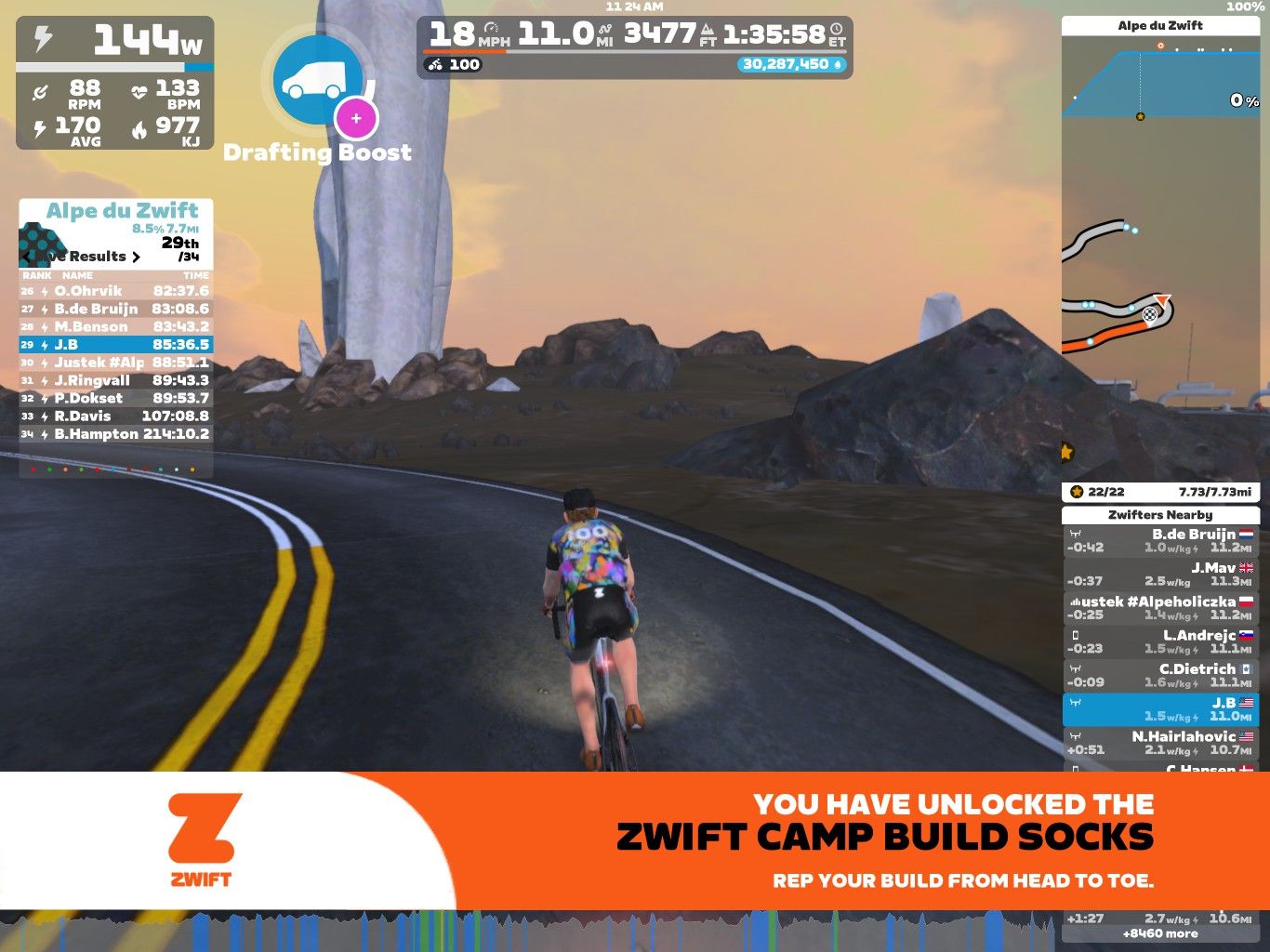 A cyclist in a virtual environment, riding on a winding road with a scenic background, representing the Zwift platform. The image displays performance stats, including power output and heart rate, as well as a leaderboard with participant names and times. A promotional banner for Zwift Camp Build Socks is shown at the bottom.