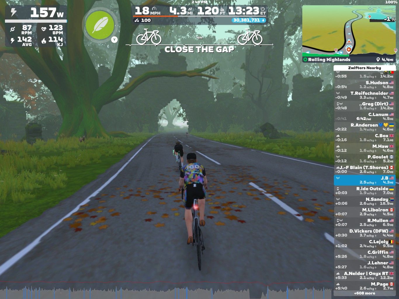 A digital cycling scene from Zwift shows three virtual cyclists riding on a leaf-strewn road surrounded by large trees. The interface displays various cycling statistics, including power output and pace, along with a map and a list of nearby users. The environment is lush and green, with a misty ambiance.