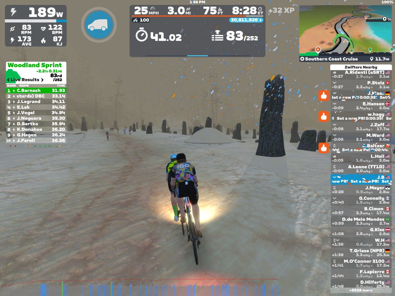 A cyclist participates in a virtual ride on Zwift, navigating a snowy landscape with large stone formations in the background. The screen displays live race results, performance metrics including speed and power output, and details of other nearby riders. The environment shows falling snow and a muted, foggy atmosphere.