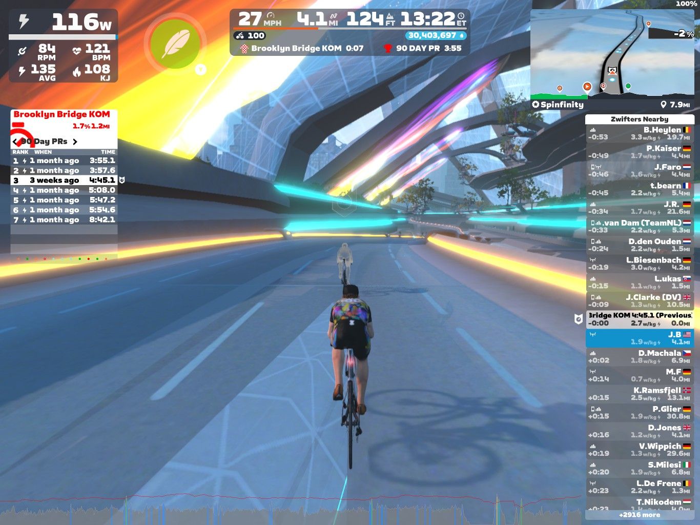 A cyclist rides on a digital road in Zwift, with colorful neon lights illuminating the space. The screen shows cycling stats, including power output, speed, and the cyclist's performance history on the Brooklyn Bridge segment. A list of nearby riders is displayed on the right side, indicating their power output and speeds. The virtual environment features futuristic elements and trees along the path.