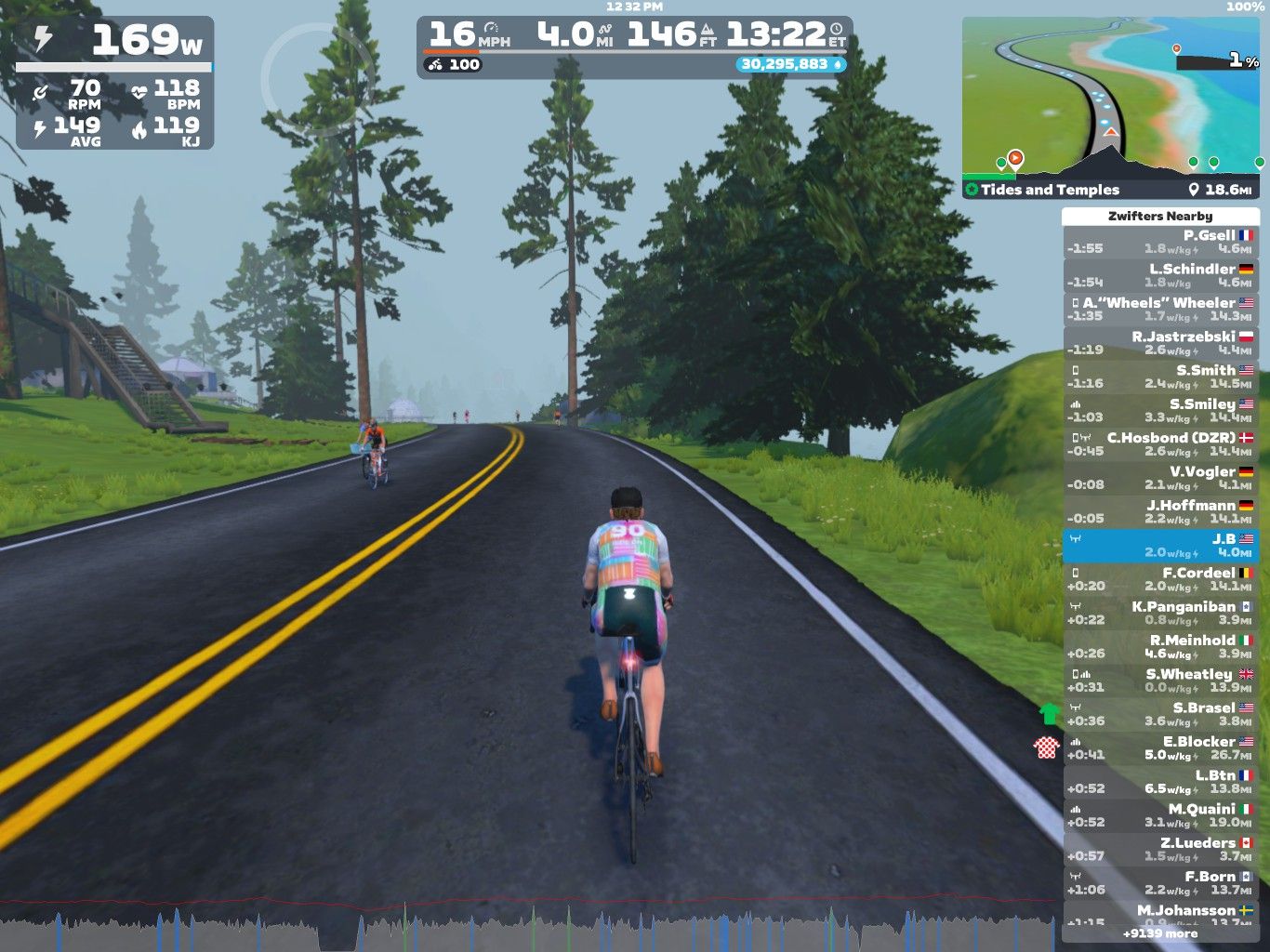 A cyclist on a virtual training course in Zwift rides along a winding road surrounded by trees. Another cyclist is visible ahead. The screen displays performance metrics, including power output, speed, distance, and heart rate, along with a leaderboard of nearby riders.