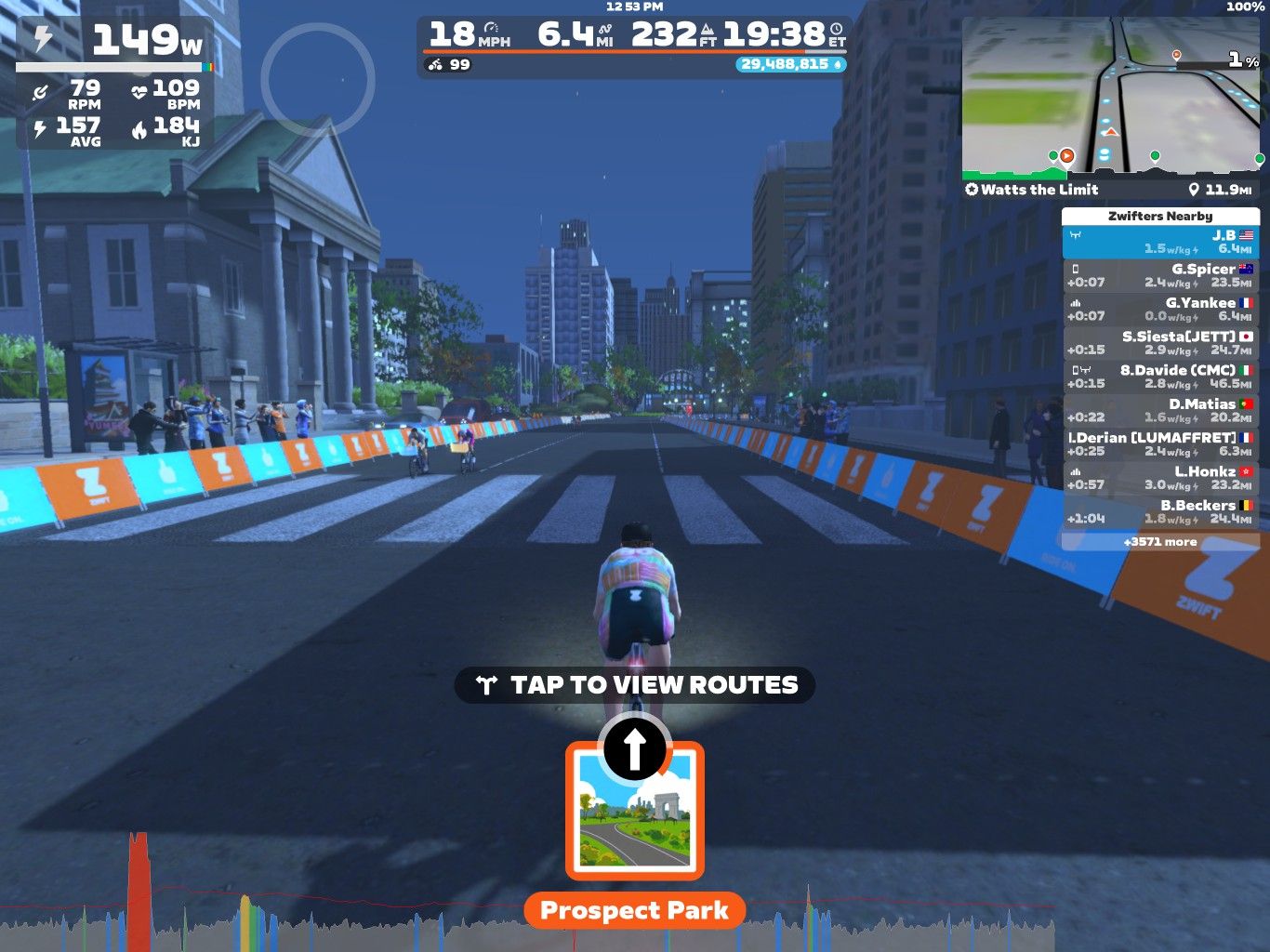 A cyclist in a virtual environment is riding down a city street in Zwift. The scene features tall buildings, a crowd of spectators, and a progress bar displaying cycling stats, including wattage, speed, and distance. The current route is labeled 