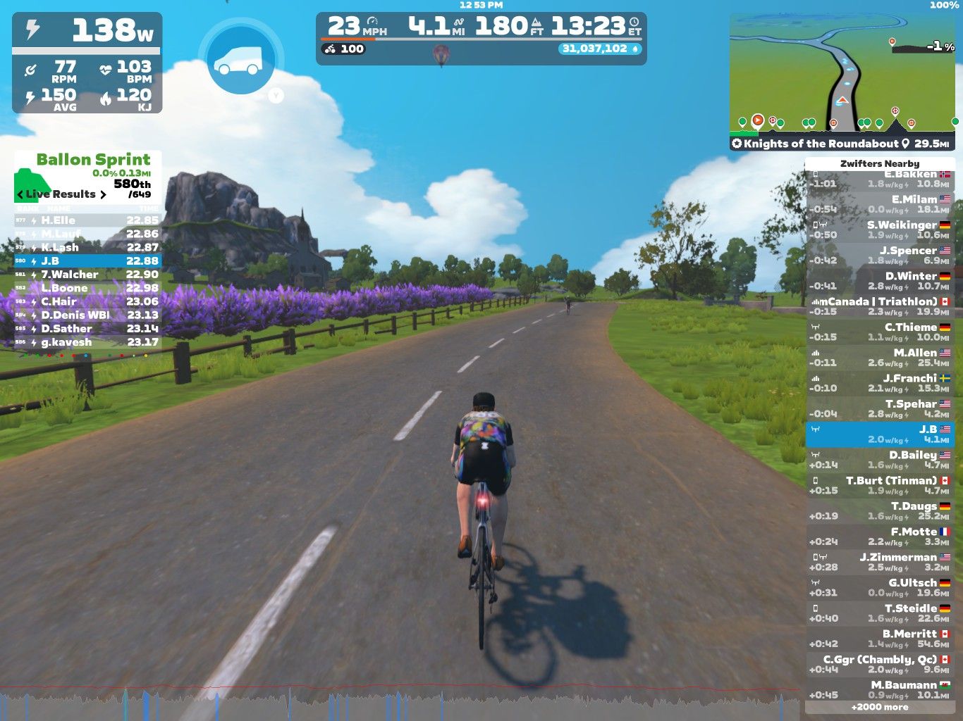 A cyclist is riding on a virtual road in Zwift, with visible metrics on power, speed, and heart rate displayed at the top. Lavender flowers line the sides of the road, and nearby hills are visible under a blue sky with white clouds. The cyclist is participating in a race, with live results and other competitors listed on the right side of the screen.