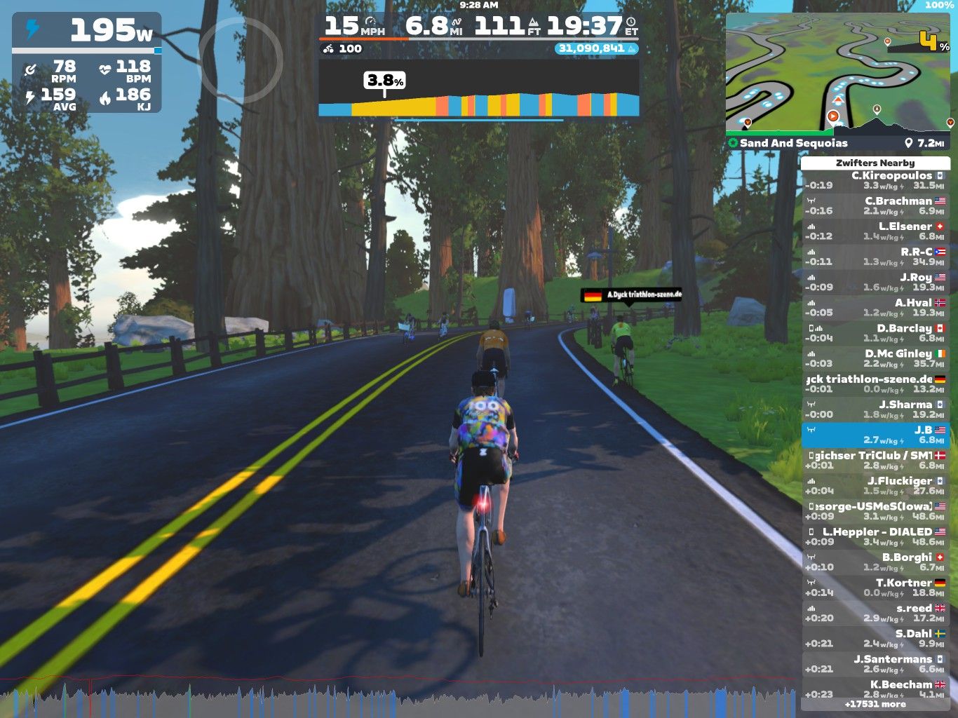 A cyclist is riding on a digital road in the Zwift virtual environment, surrounded by tall trees and a clear sky. The screen displays various performance metrics, including power output, speed, and heart rate, along with a live leaderboard and nearby cyclists. The road has two yellow lines and curves ahead.
