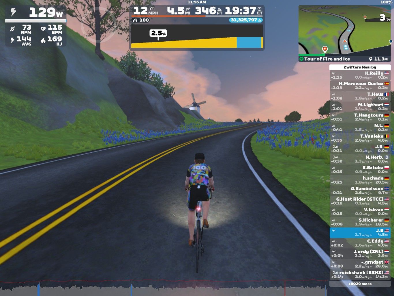A cyclist rides on a virtual road in Zwift, surrounded by green hills and purple flowers. The cyclist has a colorful outfit and the screen displays performance metrics, including speed, distance, and heart rate. The sky in the background shows a sunset. A leaderboard on the right lists other riders nearby.
