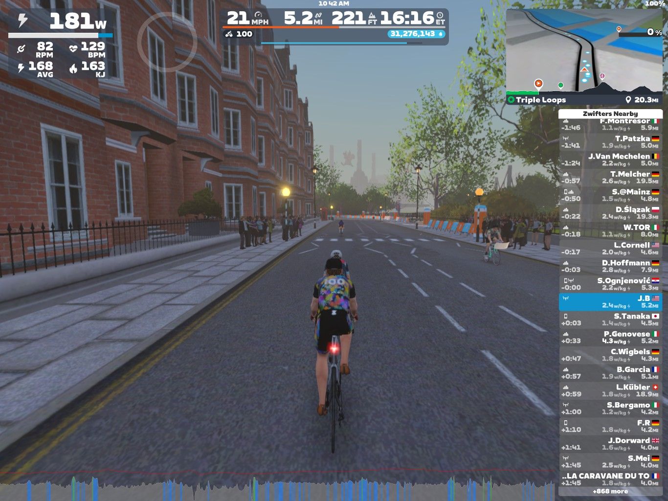 A cyclist is riding on a virtual street in Zwift, surrounded by buildings and trees. The cyclist is wearing a colorful jersey and there are several virtual spectators along the roadside. A scoreboard on the side displays cycling stats and nearby competitors.