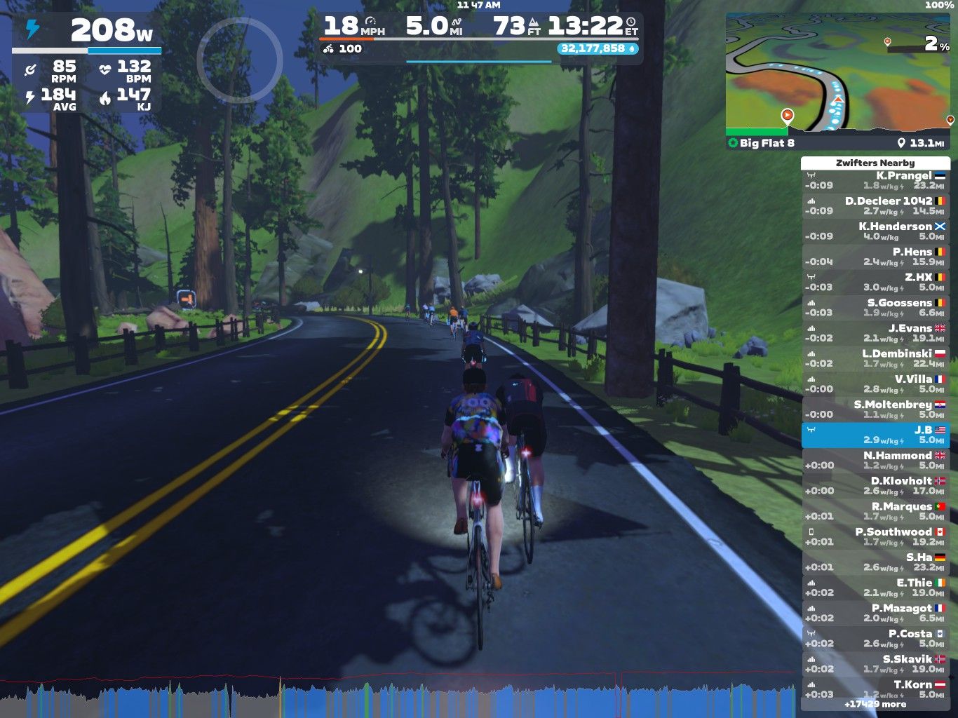 A group of cyclists riding on a virtual road in a scenic landscape, with tall trees and mountains in the background. The screen displays various statistics like power output, speed, and heart rate, along with a list of nearby cyclists.