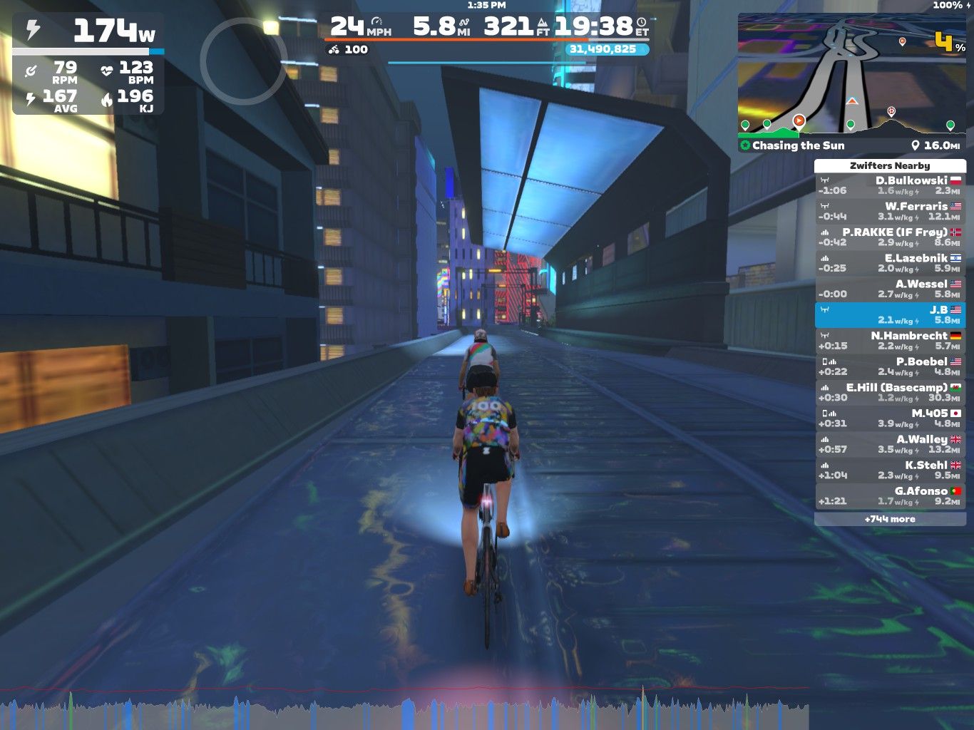 A cyclist rides through a virtual cityscape in Zwift, displaying speed, power, and heart rate metrics on the screen. The environment features bright lights and modern buildings, with a leaderboard showing nearby cyclists and their metrics. The cyclist is wearing a colorful jersey and is lit up by virtual effects as they pedal along the illuminated path.