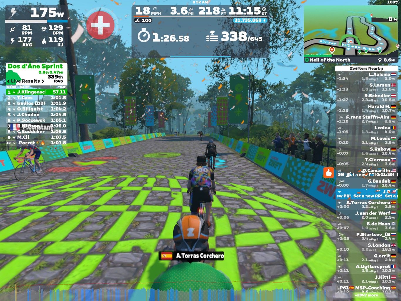 A cycling race is taking place in a virtual environment on Zwift, featuring riders on a colorful, stylized road with green graphics. A leaderboard displays live results, with information about speed, heart rate, and distance. In the background, trees and banners are visible, and the atmosphere is festive with visual effects.
