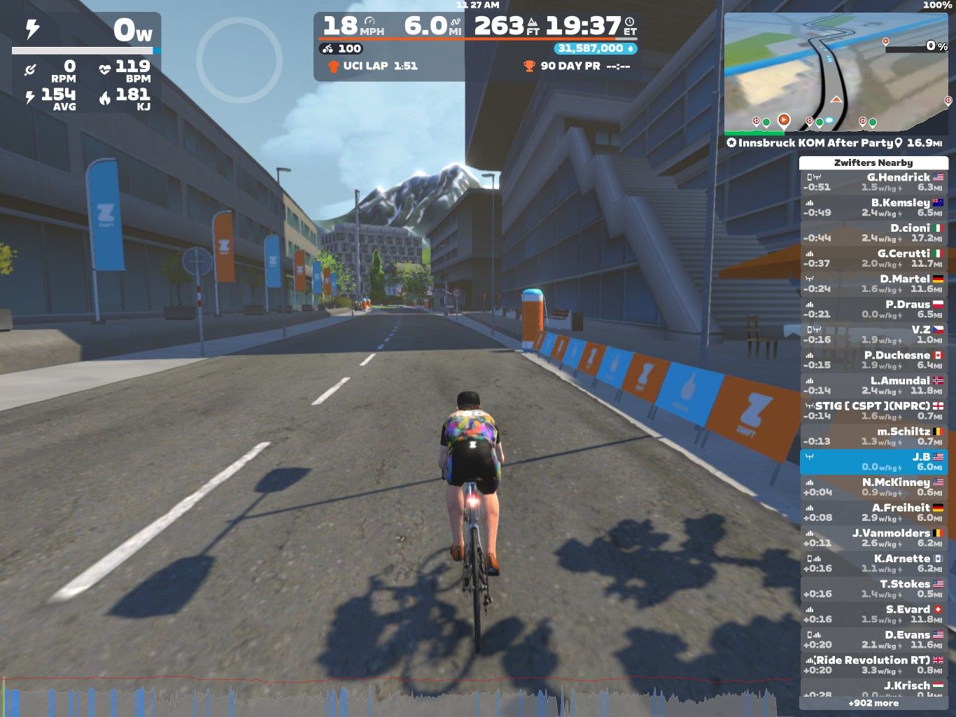 A cyclist rides along a virtual road in Zwift, surrounded by buildings and banners. The screen displays stats such as speed (18 mph), distance (6.0 miles), and other performance metrics, along with a leaderboard of nearby participants. The environment features trees, mountains in the background, and a digital interface showing the course details.