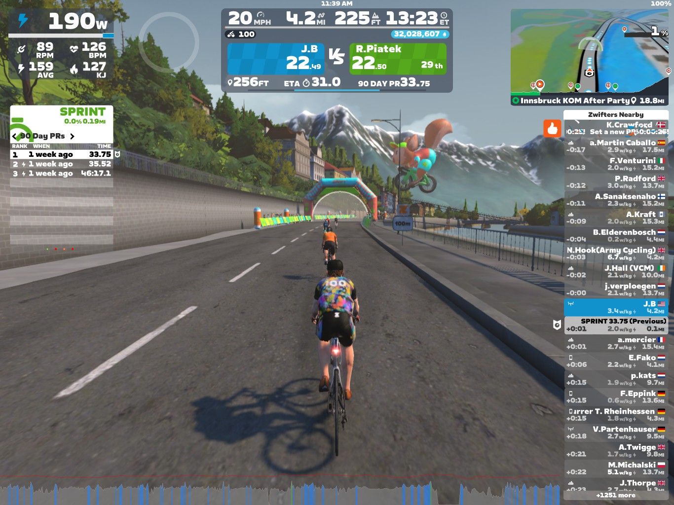 A digital cycling scene in Zwift featuring three cyclists on a road. The cyclists are riding towards a colorful archway, with a mountain backdrop. The screen displays various statistics about power, speed, distance, and nearby users on the platform.