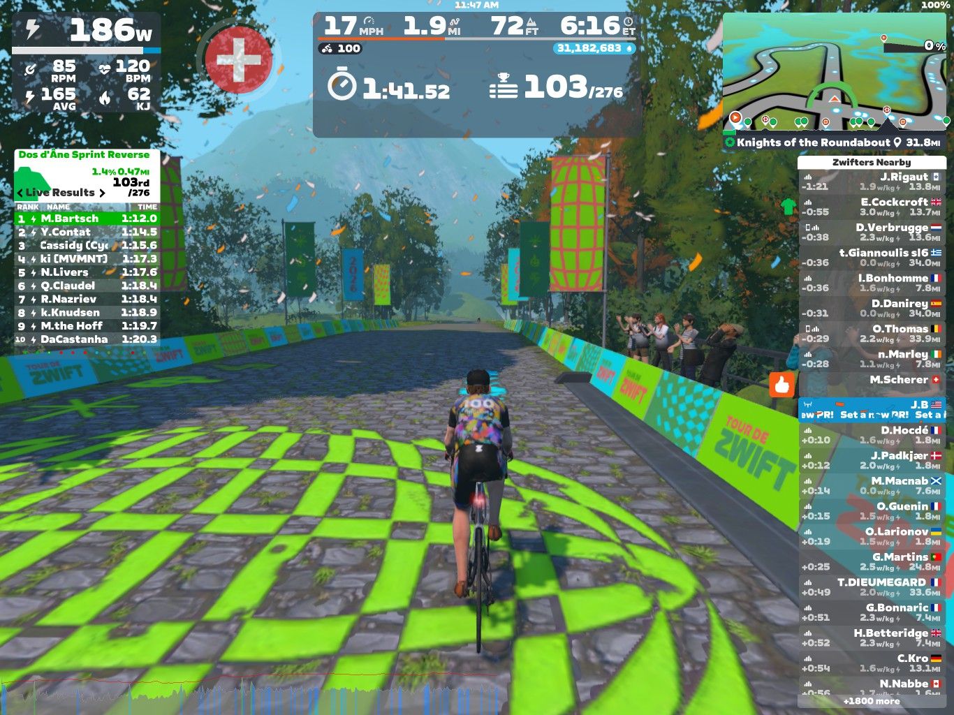 A cyclist rides on a virtual course in Zwift, displayed on a digital screen. The scene shows a cobblestone path lined with colorful banners, with trees and mountains in the background. The cyclist's stats are visible, including speed, distance, and power output, along with a leaderboard showing real-time results of competitors. The atmosphere is lively, with cheering spectators depicted along the route.