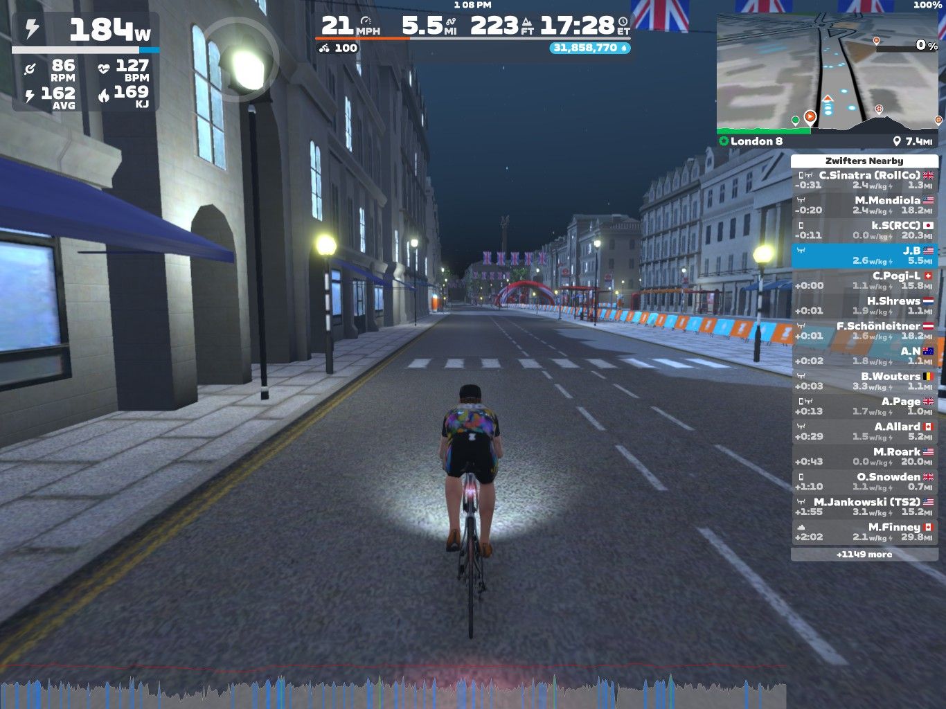 A virtual cycling scene in Zwift showing a rider on a street at night, surrounded by buildings with illuminated streetlights. The interface displays cycling stats including power output, speed, and distance, along with a map of the route labeled 
