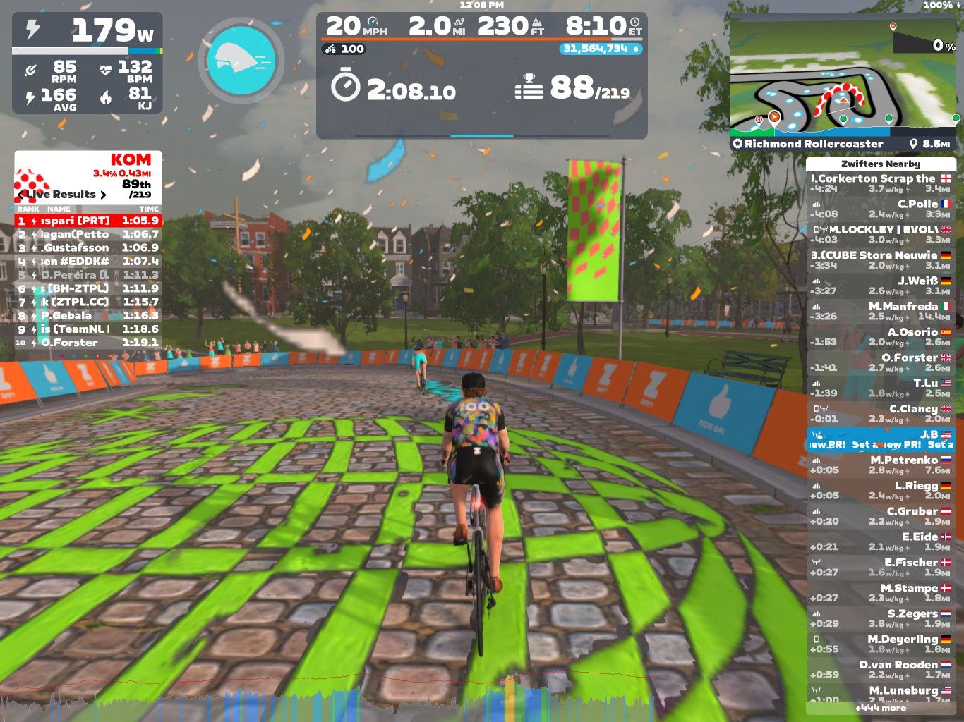 A cyclist navigates a colorful virtual course in Zwift, showing speed and power metrics on the screen. In the background, people are cheering, and the course features green and striped patterns along the cobblestone path. Various performance statistics and rankings are displayed on the side of the screen.