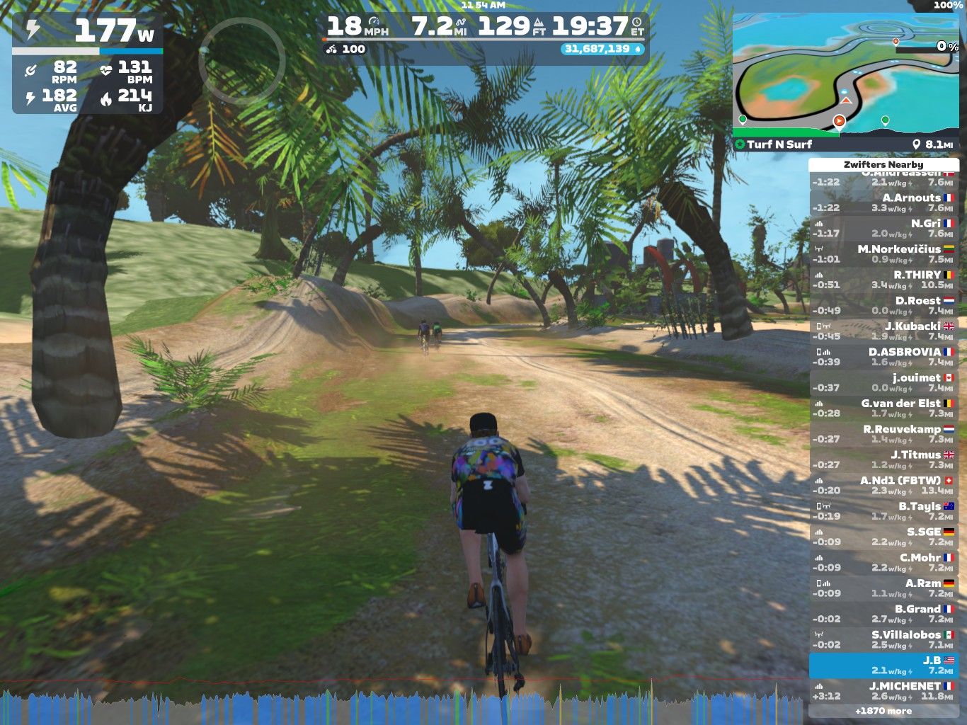 A cyclist rides on a dirt path surrounded by trees in a virtual environment, showing power and speed metrics. The screen displays current stats including power in watts, heart rate, and miles cycled, along with a list of nearby cyclists and their performance times. A mini-map is visible in the top right corner.