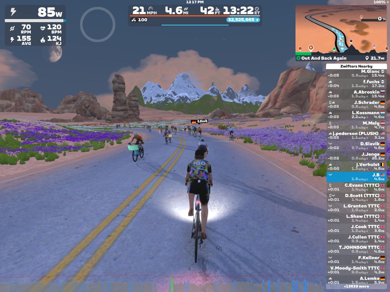 A screenshot from Zwift shows several cyclists riding on a road surrounded by colorful flowers and rocky mountains. The display includes cycling statistics such as wattage, speed, and distance, along with a list of nearby riders. The setting features a clear sky with a visible moon.