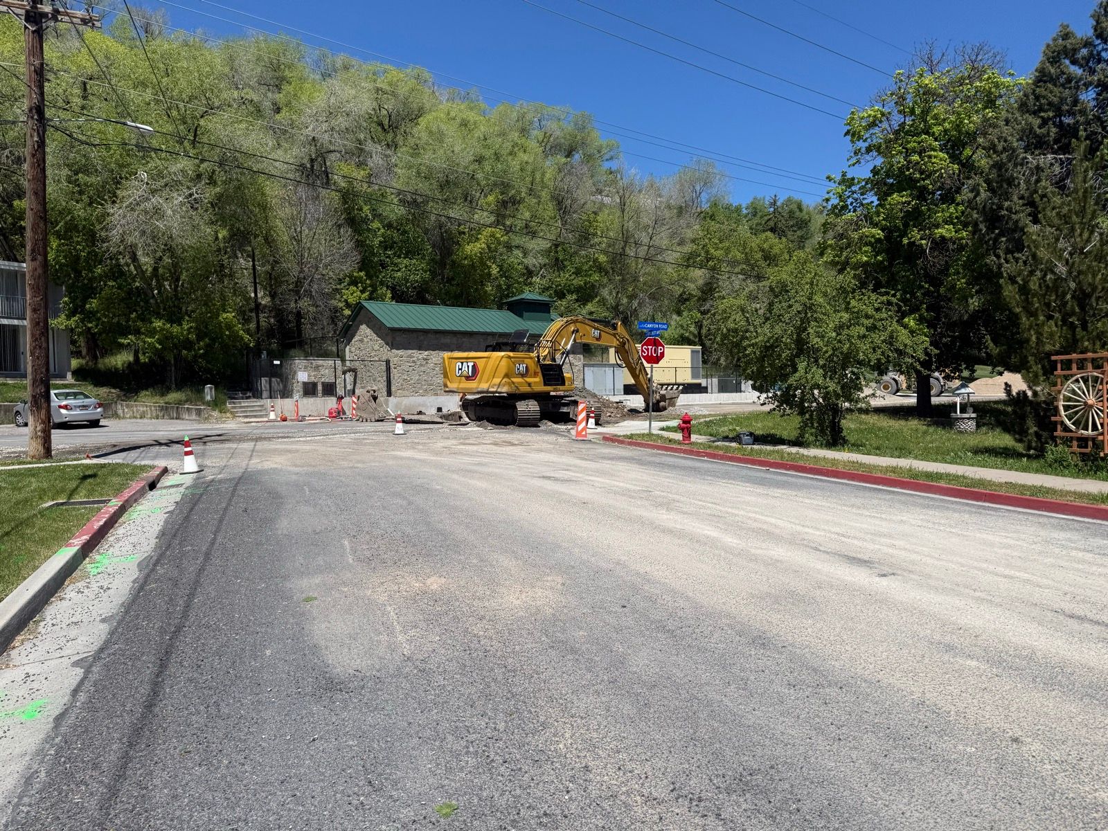 A construction site in Paradise, Utah, featuring a large yellow excavator parked on a newly laid asphalt road. There are traffic cones and a 