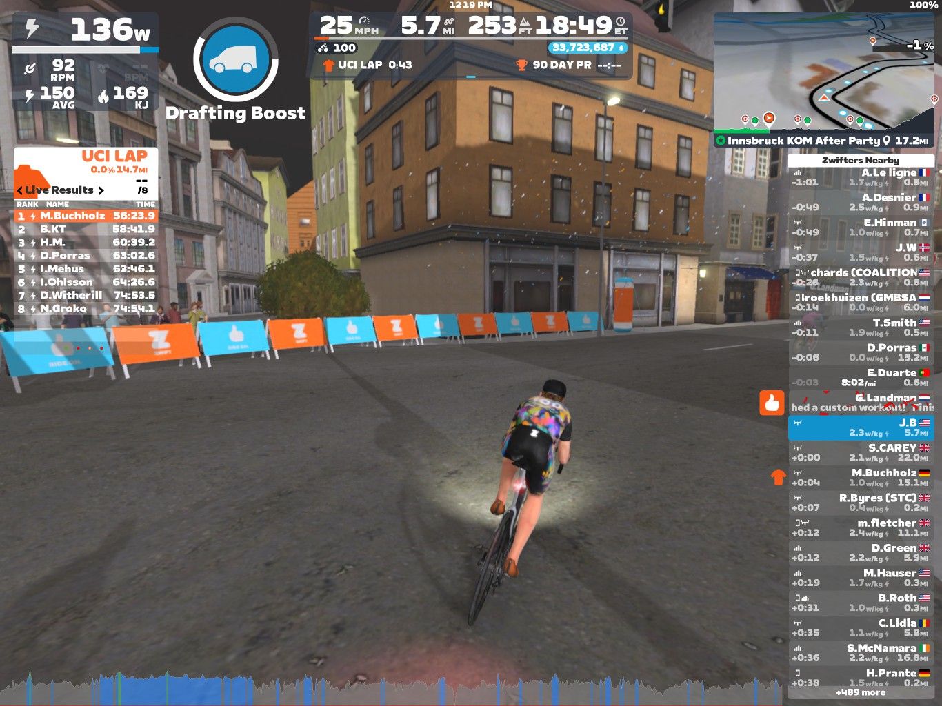 A cyclist is riding on a virtual course in Zwift, wearing a colorful jersey. The screen displays various performance metrics, including power output, speed, and lap times. In the background, there are buildings and barriers marked with the Zwift logo, indicating a competitive cycling environment.