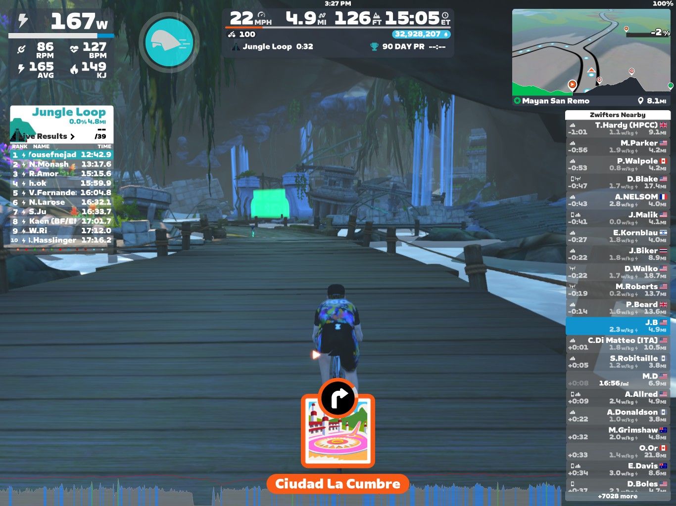 A screenshot from Zwift showing a virtual cycling environment on the Jungle Loop. The cyclist is traveling on a wooden bridge surrounded by green trees and rock formations. The screen displays various statistics, including speed, power output, and heart rate. Rankings and results for multiple cyclists are shown on the left side, while nearby Zwifters and a map are visible on the right. The location is labeled 