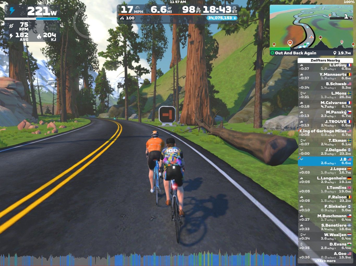 Two cyclists riding on a virtual road in Zwift's simulated environment, surrounded by trees and mountains. The screen displays their current speed, wattage, and other cycling stats, along with a mini-map and names of nearby riders.