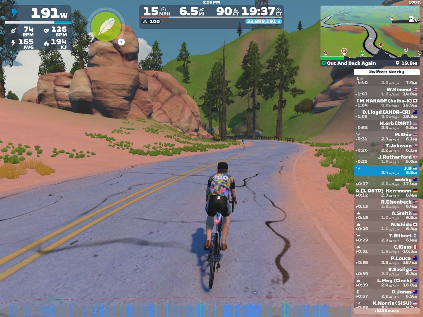 A cyclist wearing a colorful jersey rides on a virtual road in Zwift, surrounded by green trees and rock formations. The screen displays performance metrics, including wattage, speed, heart rate, and distance traveled.