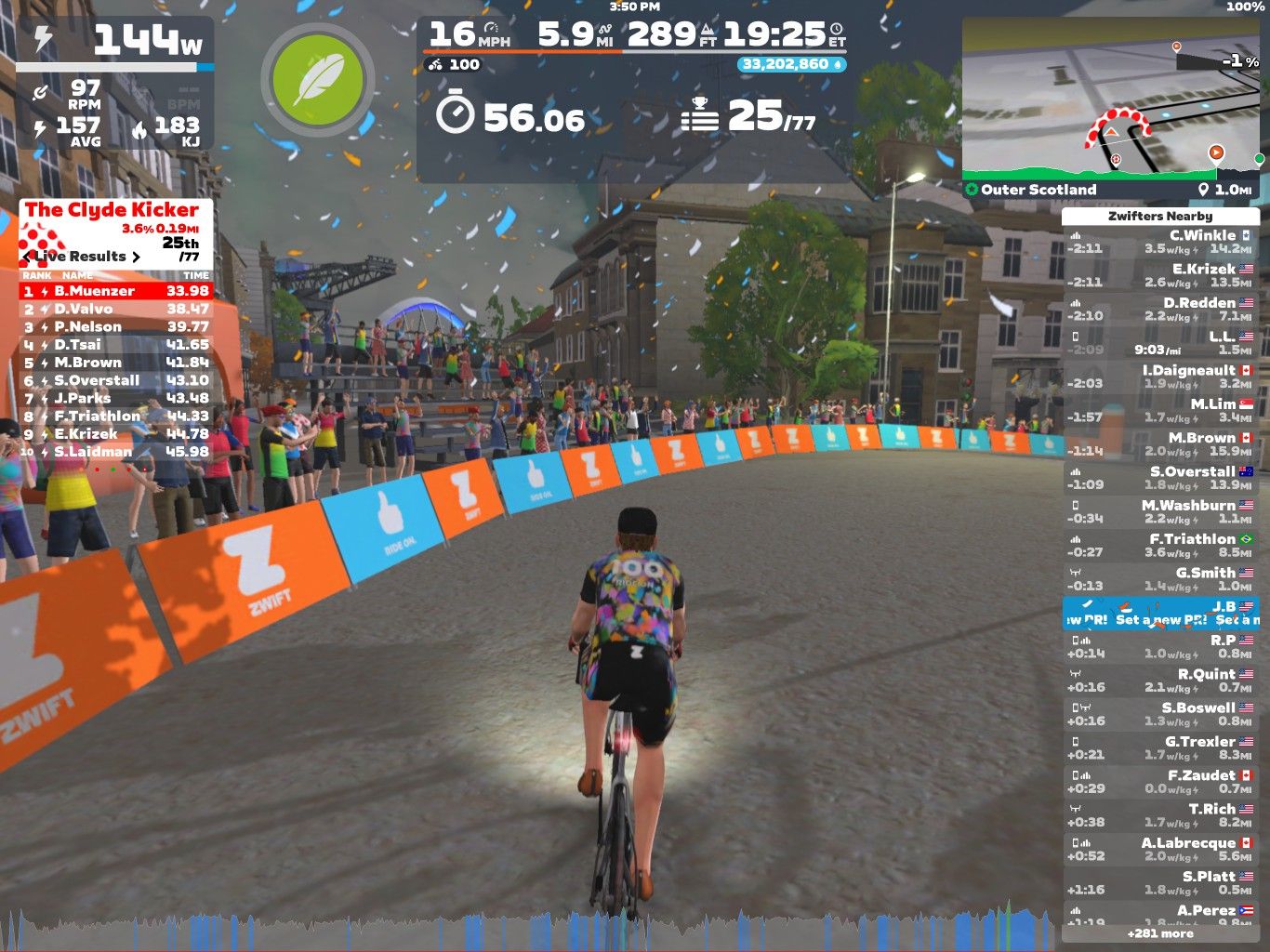A cyclist rides on a virtual track in Zwift, surrounded by spectators cheering on the sidelines. The cyclist is referencing live race results displayed on the screen, showing rankings and times for participants. The background features colorful banners and elements typical of an outdoor racing environment, while confetti falls from above, adding to the festive atmosphere.