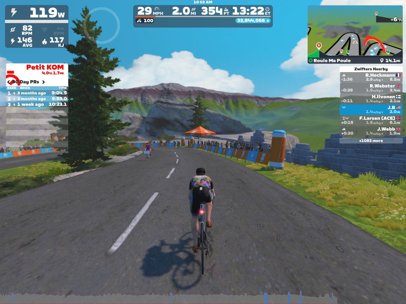 A cyclist rides along a virtual road in Zwift, surrounded by greenery and mountains in the background. The screen displays performance metrics, including speed and power, as well as information about nearby cyclists.