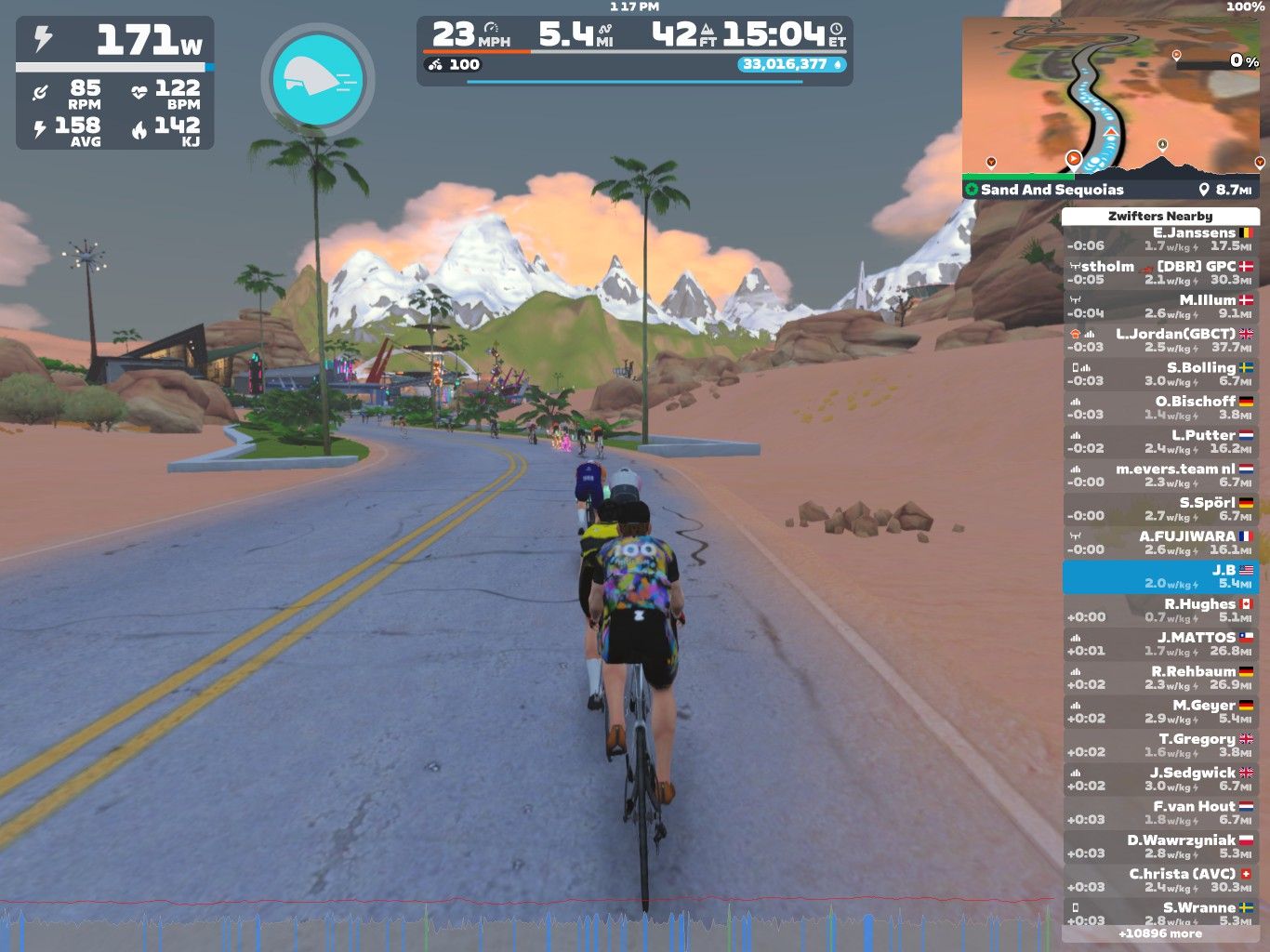 A virtual cycling scene in Zwift, showing cyclists riding on a road with palm trees and mountains in the background. The cyclists are near a turn, and a leaderboard displays nearby riders' names and statistics. The interface shows various performance metrics like power, speed, and distance.
