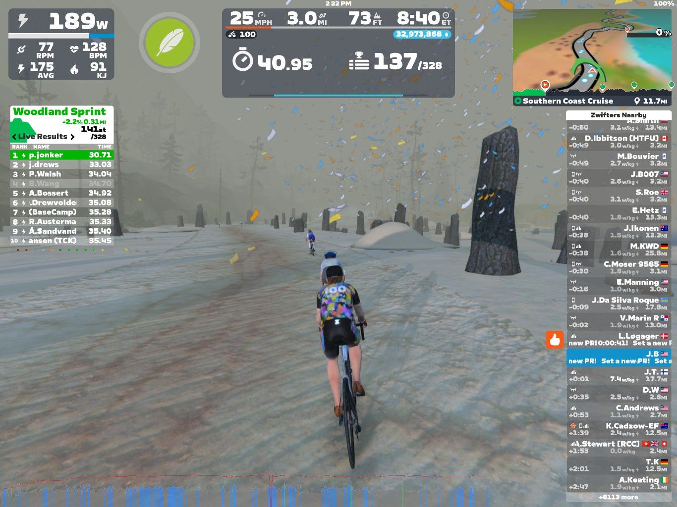 A cyclist rides on a snowy path in Zwift, surrounded by tall stone markers. The screen displays performance metrics, including watts, speed, and heart rate, alongside a leaderboard showing live results of a sprint event.