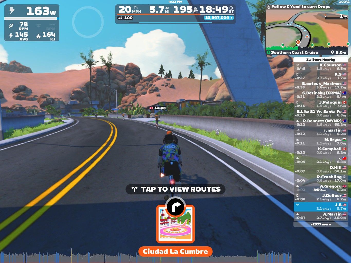 A virtual cycling scene from Zwift, showing a bike rider on a smooth, winding road. The landscape features rocky hills and greenery under a clear blue sky. On-screen stats display power, speed, and distance, along with nearby cyclists’ names and times. A route selection button is visible at the bottom. The location is labeled 