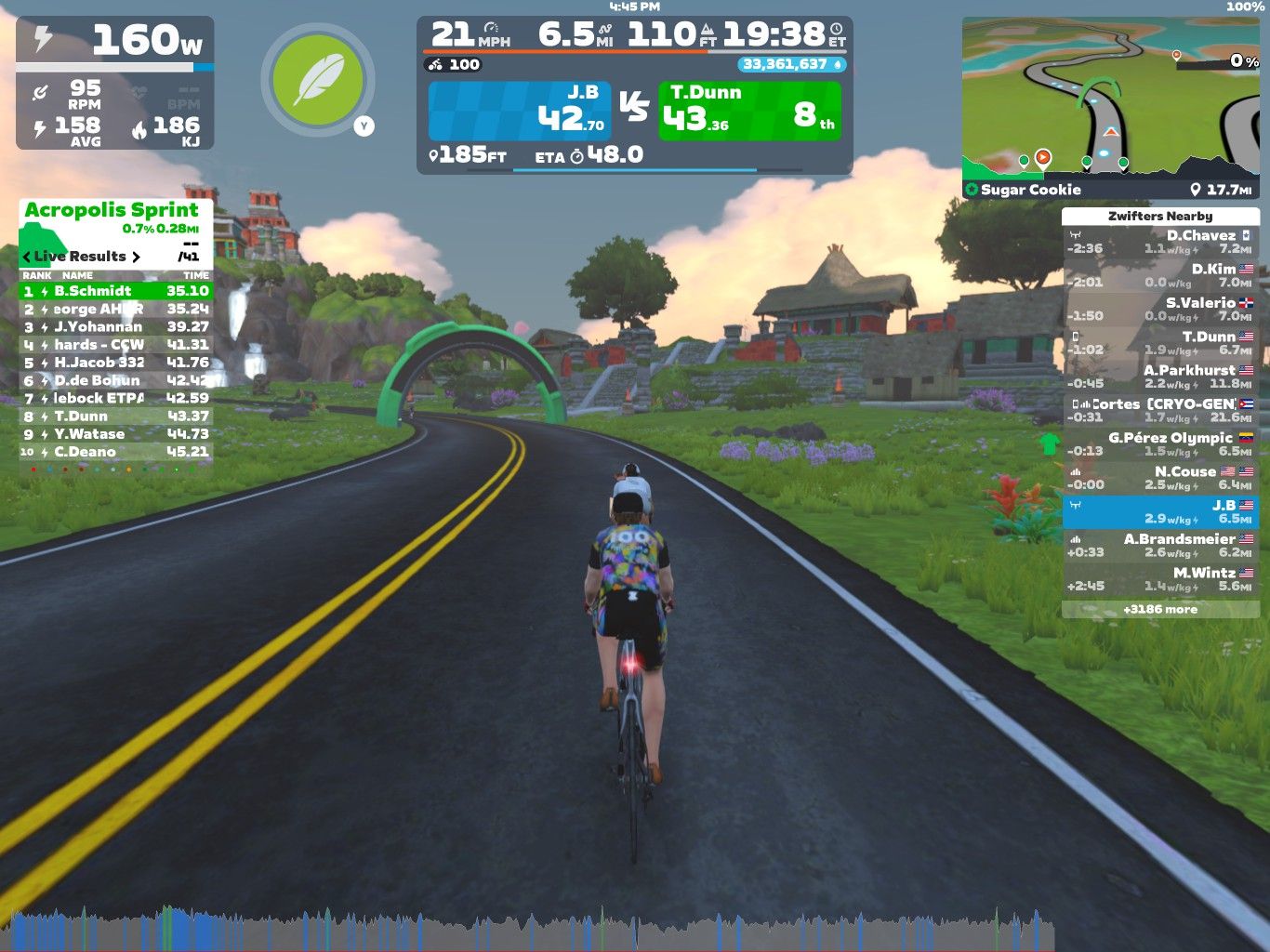 A cyclist participates in a virtual race on Zwift, riding on a winding road in a colorful, cartoon-like landscape. The display shows various statistics, including power output, speed, and race standings, with surrounding scenery featuring hills, trees, and buildings.