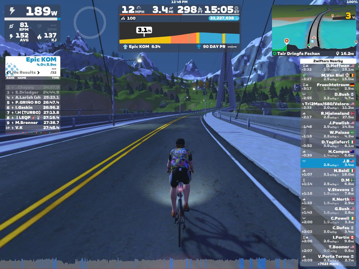 A cyclist is riding on a virtual road with mountains in the background, part of the Zwift platform. The screen displays various performance metrics, including power output, speed, distance, and a leaderboard with rankings and times. There is also a map indicating the cyclist's location within the virtual environment.