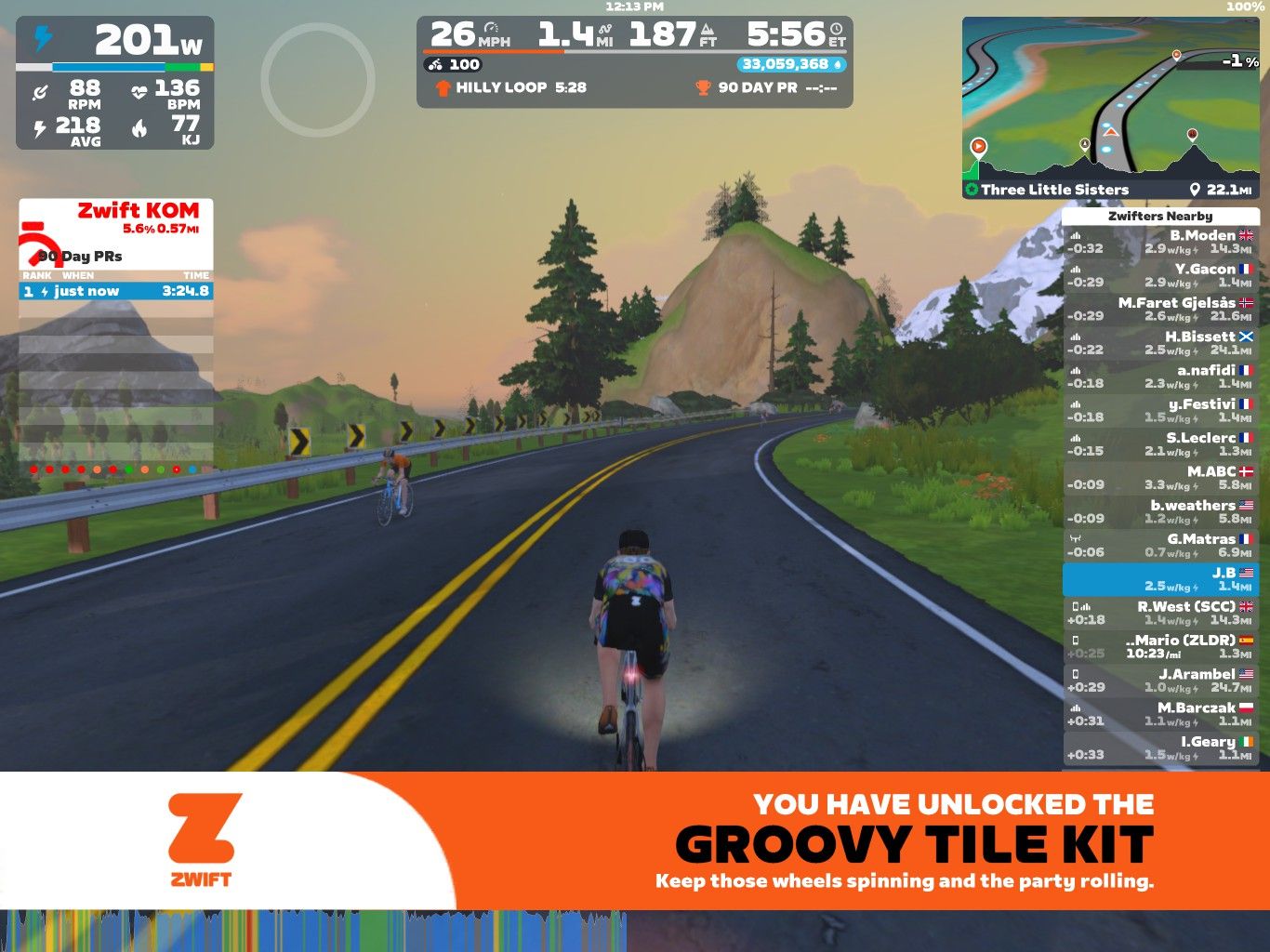 A screenshot from Zwift, an online cycling platform. The scene shows two virtual cyclists riding on a winding road with mountains and trees in the background. Important statistics such as power output, speed, and heart rate are displayed on the screen, along with leaderboard information and a notification about unlocking a new item.
