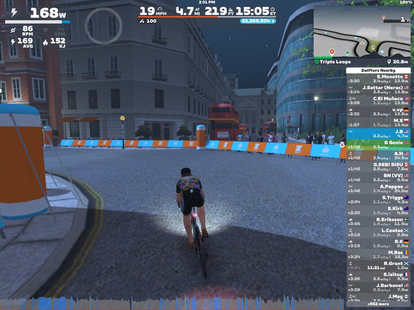 A cyclist is riding on a virtual course in Zwift during a nighttime setting. The cyclist is shown from behind, with a focus on stats like power output and speed displayed on the screen. Surrounding the cyclist are virtual buildings, a road lined with barriers, and a map indicating the course layout. Several other participants are visible nearby, and a double-decker bus can be seen in the background.