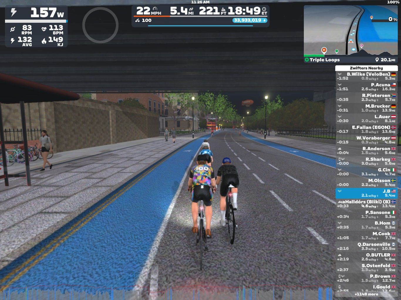 A digital cycling scene in Zwift, featuring cyclists riding on a blue bike lane under an overpass. The setting includes a path with bikes parked nearby, city buildings in the background, and a visible leaderboard displaying nearby riders' stats and times. The time on the clock shows 18:49.