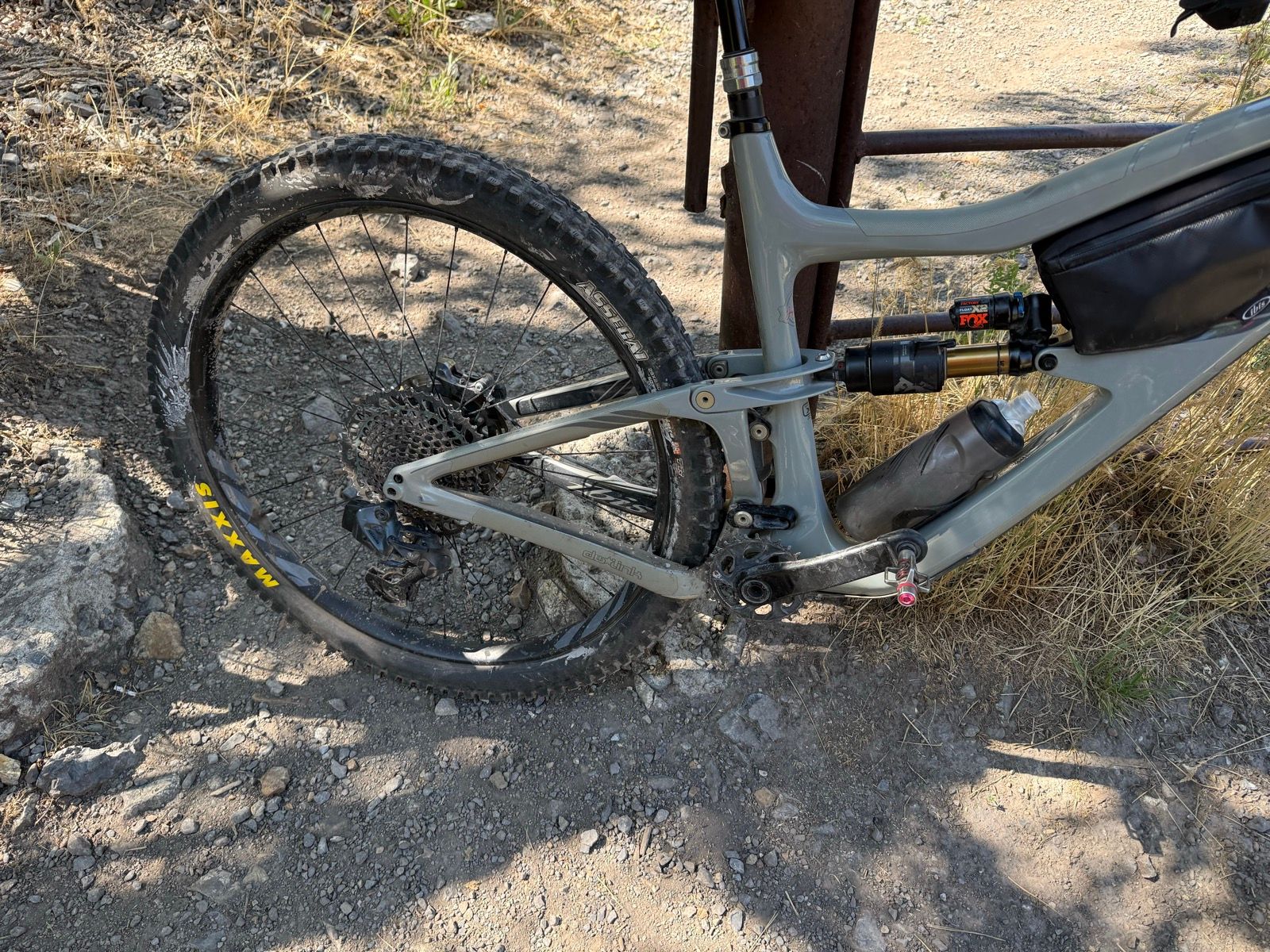 A close-up view of a mountain bike resting on a gravel path in Providence Canyon, Utah. The image shows the rear wheel and part of the bike's frame, highlighting the tire's treads and a water bottle attached to the bike. Surrounding the bike are patches of dry grass and small rocks.