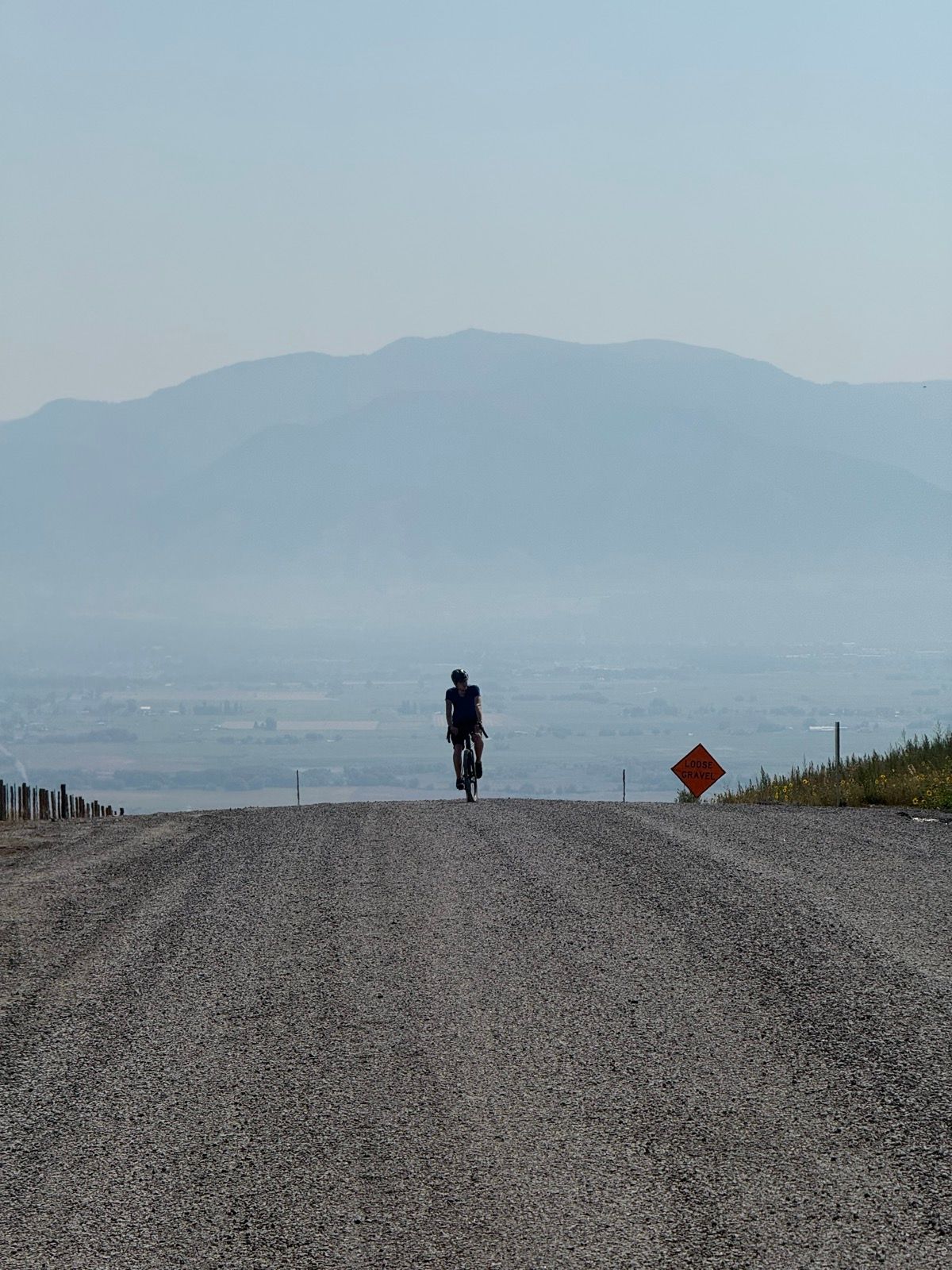 A person rides a bicycle on a gravel road leading into a hazy valley, with mountains in the background. A warning sign for loose gravel is visible on the right side. The scene is set in Logan, Utah, United States.