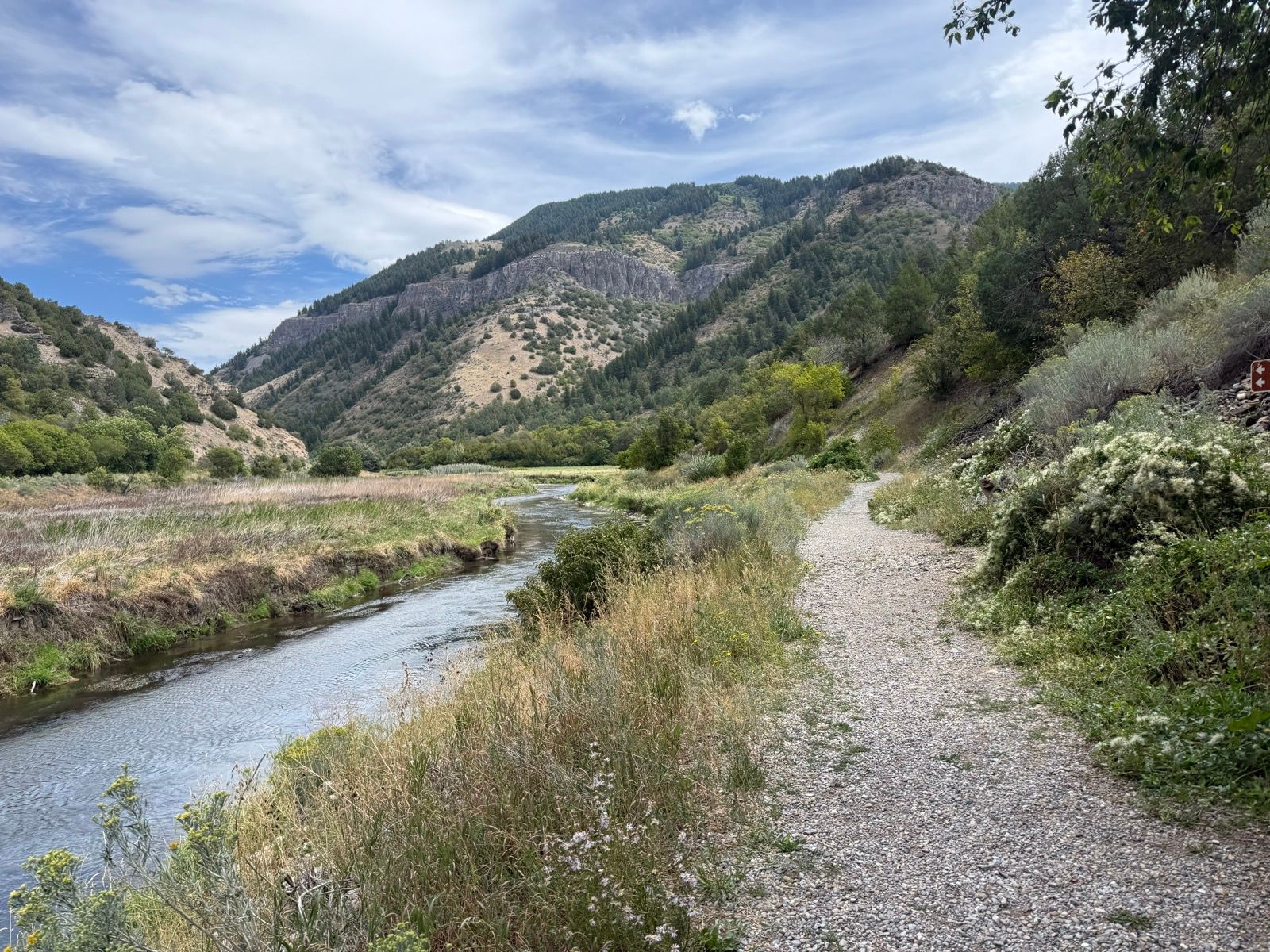 A gravel path winds through tall grass and shrubs alongside a stream, with mountainous terrain in the background under a partly cloudy sky. The scene is set in Providence, Utah, showcasing the area's natural beauty.