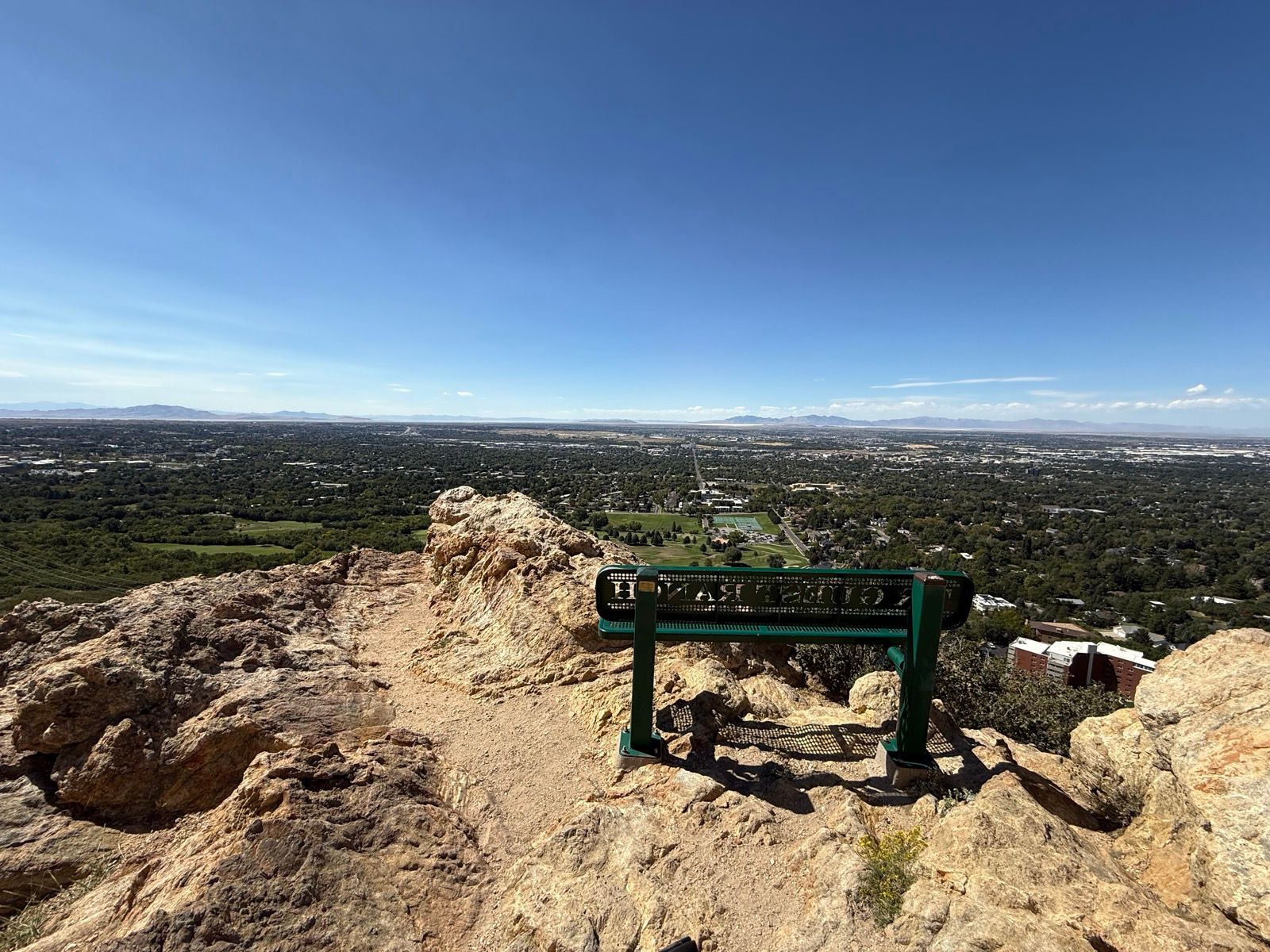 A panoramic view from a rocky viewpoint in Ogden, UT, showing a valley with green fields and city structures beneath a clear blue sky. A lookout bench is positioned at the edge of the rocks.