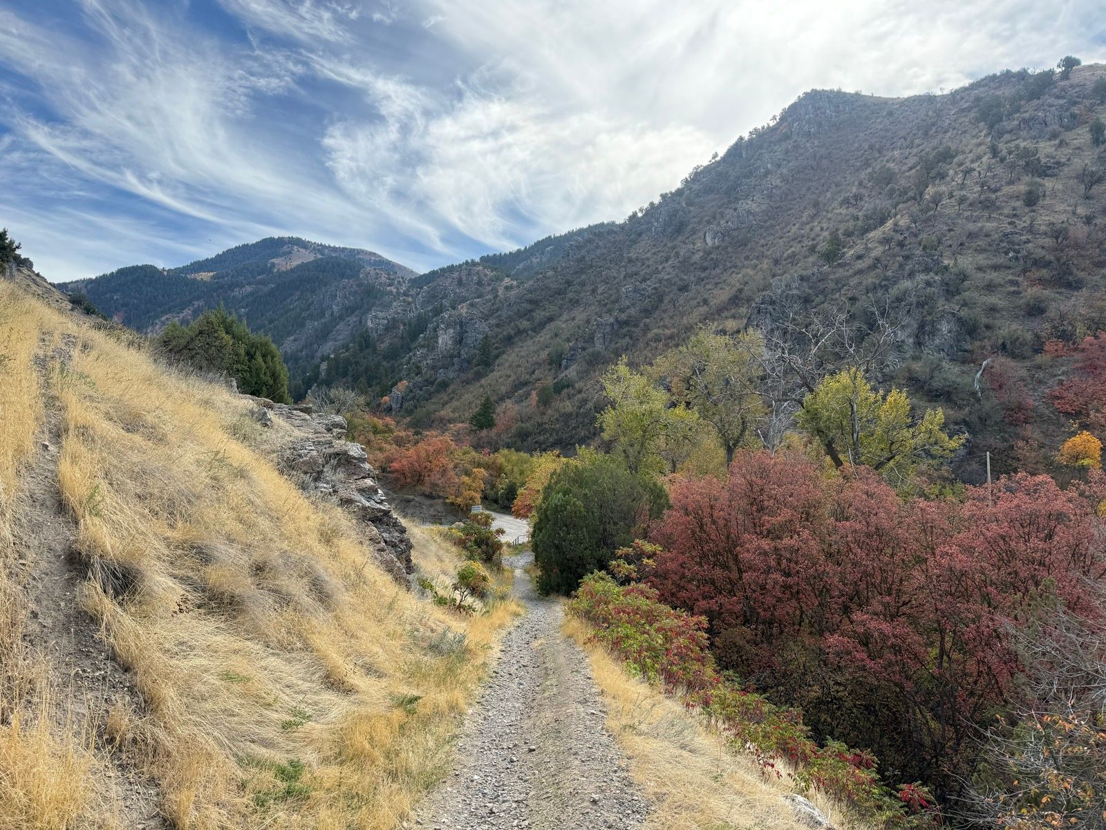 A gravel path winds through a scenic landscape at Logan River Trail, flanked by rocky hills and colorful autumn foliage. The sky is partly cloudy, adding depth to the mountainous backdrop.