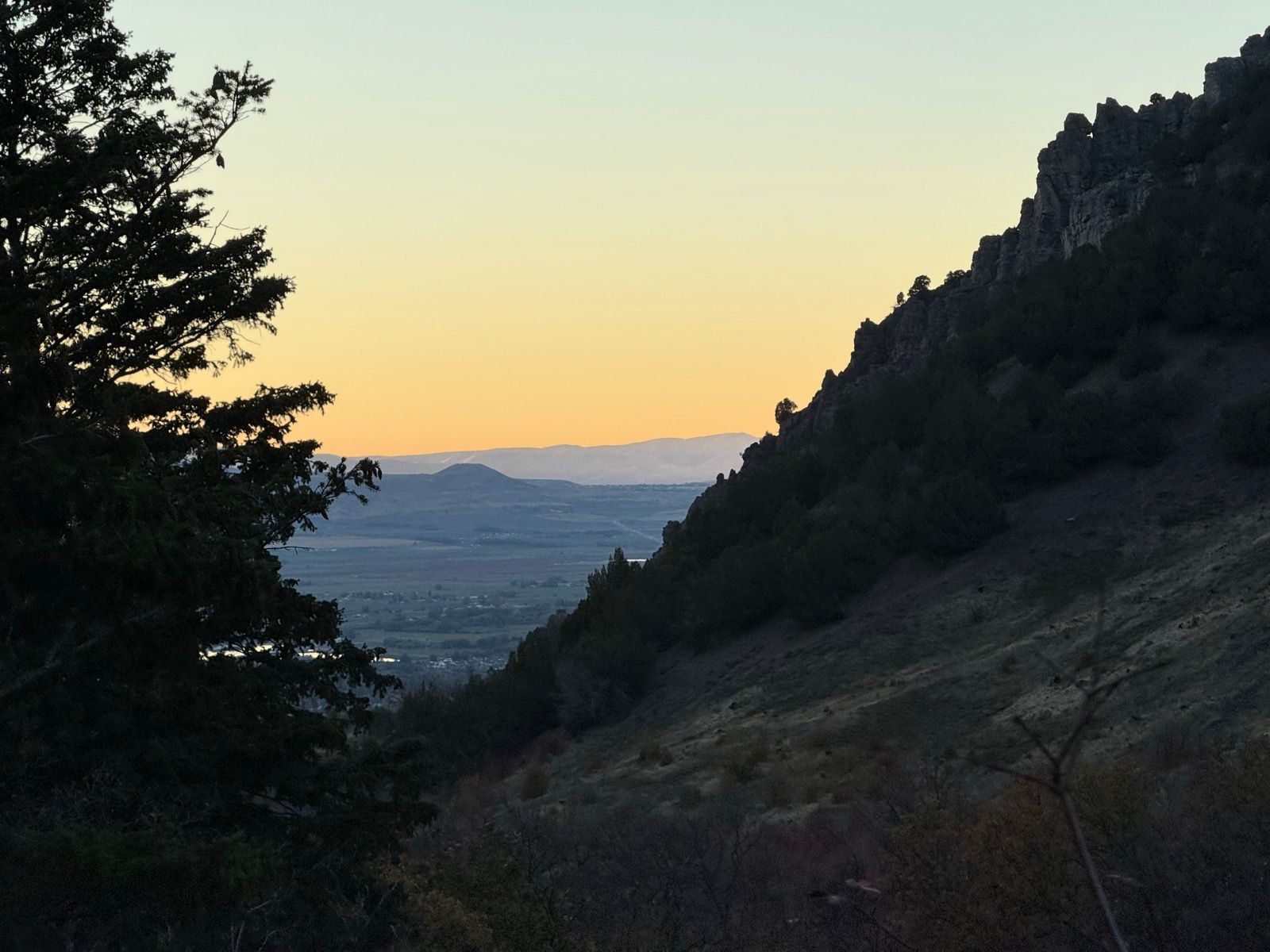 A view of Providence Canyon in Utah during sunset, featuring silhouetted mountain ridges and a valley below, with trees on the left side of the image and a soft orange and blue sky.