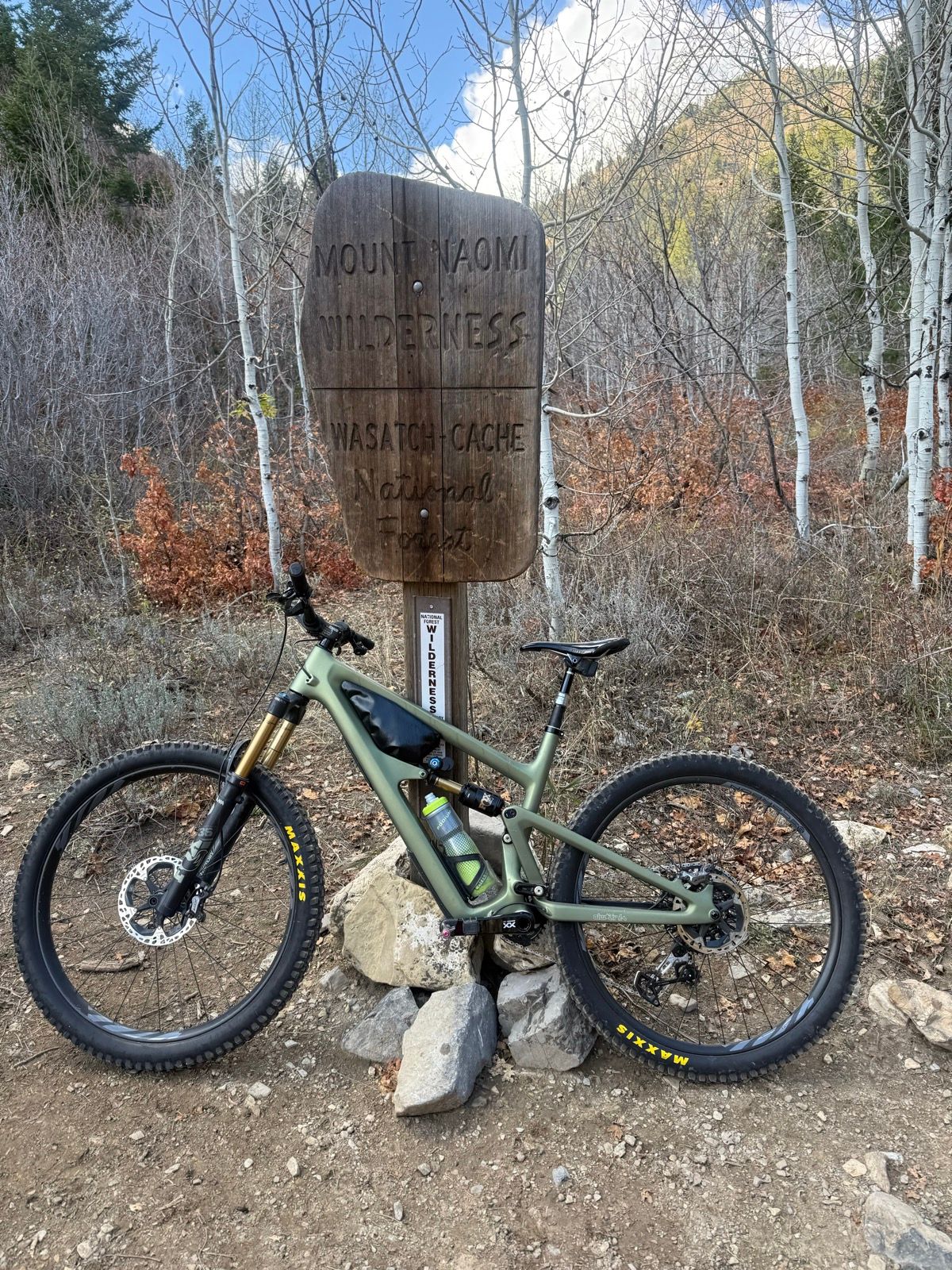A mountain bike is leaning against a wooden sign that reads "MOUNT NAOMI WILDERNESS" in the Green Canyon area of Utah. The background features sparse trees and a trail with rocky terrain.