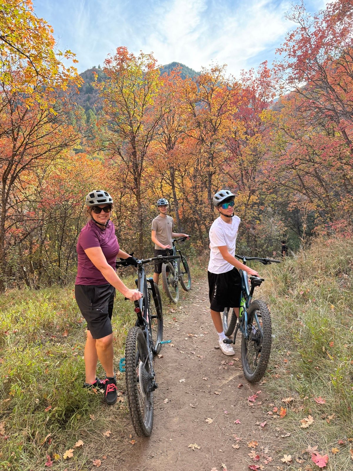 A woman and two boys are standing on a dirt path in a forested area with colorful fall foliage. They are wearing helmets and holding mountain bikes. The background features trees with vibrant orange and yellow leaves, and a blue sky with wispy clouds above.