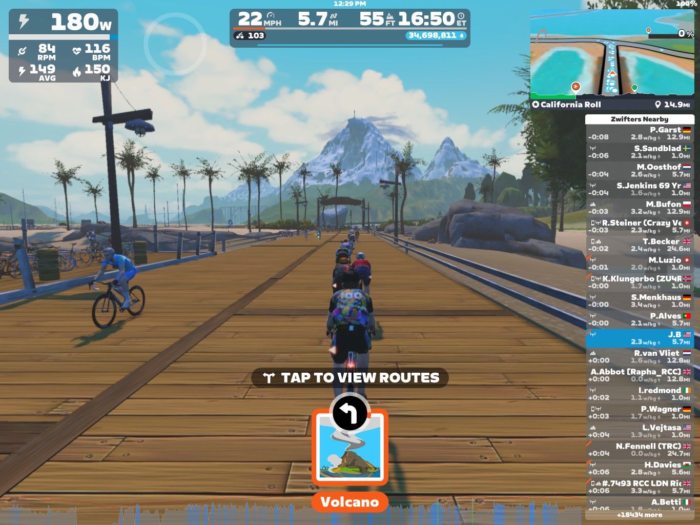 A digital cycling scene in Zwift shows several cyclists riding on a wooden pathway surrounded by palm trees and mountains. The interface displays various cycling stats, including speed and distance, along with a map showing nearby routes. A cyclist in blue leads the group, and there's a prominent focus on the "Volcano" route option.