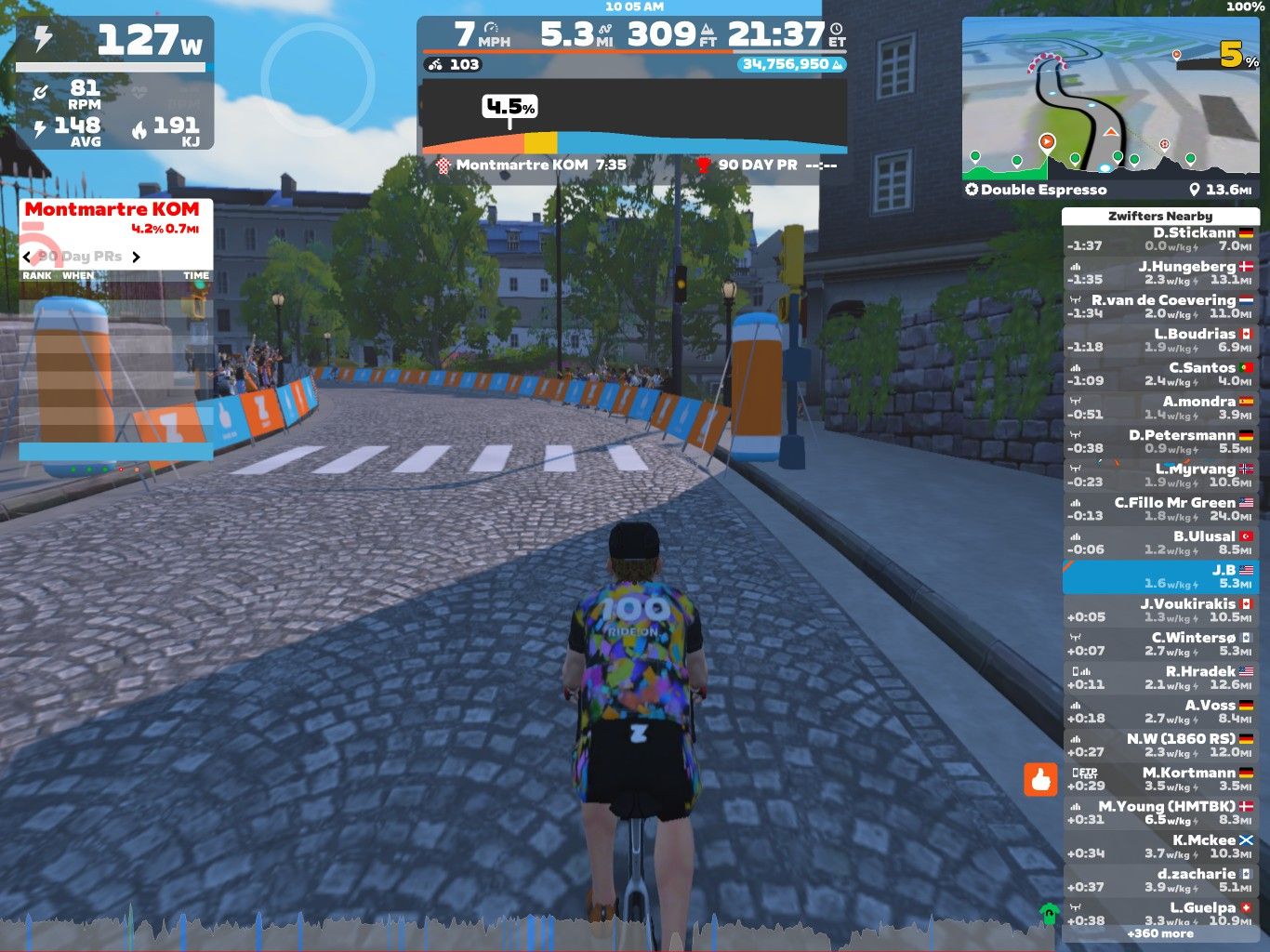 A cyclist riding on a cobblestone street in a virtual setting, displaying statistics on speed, power, and elevation. The scene features a bend in the road with orange barriers and trees lining the path, along with a leaderboard showing nearby cyclists and their performance metrics.